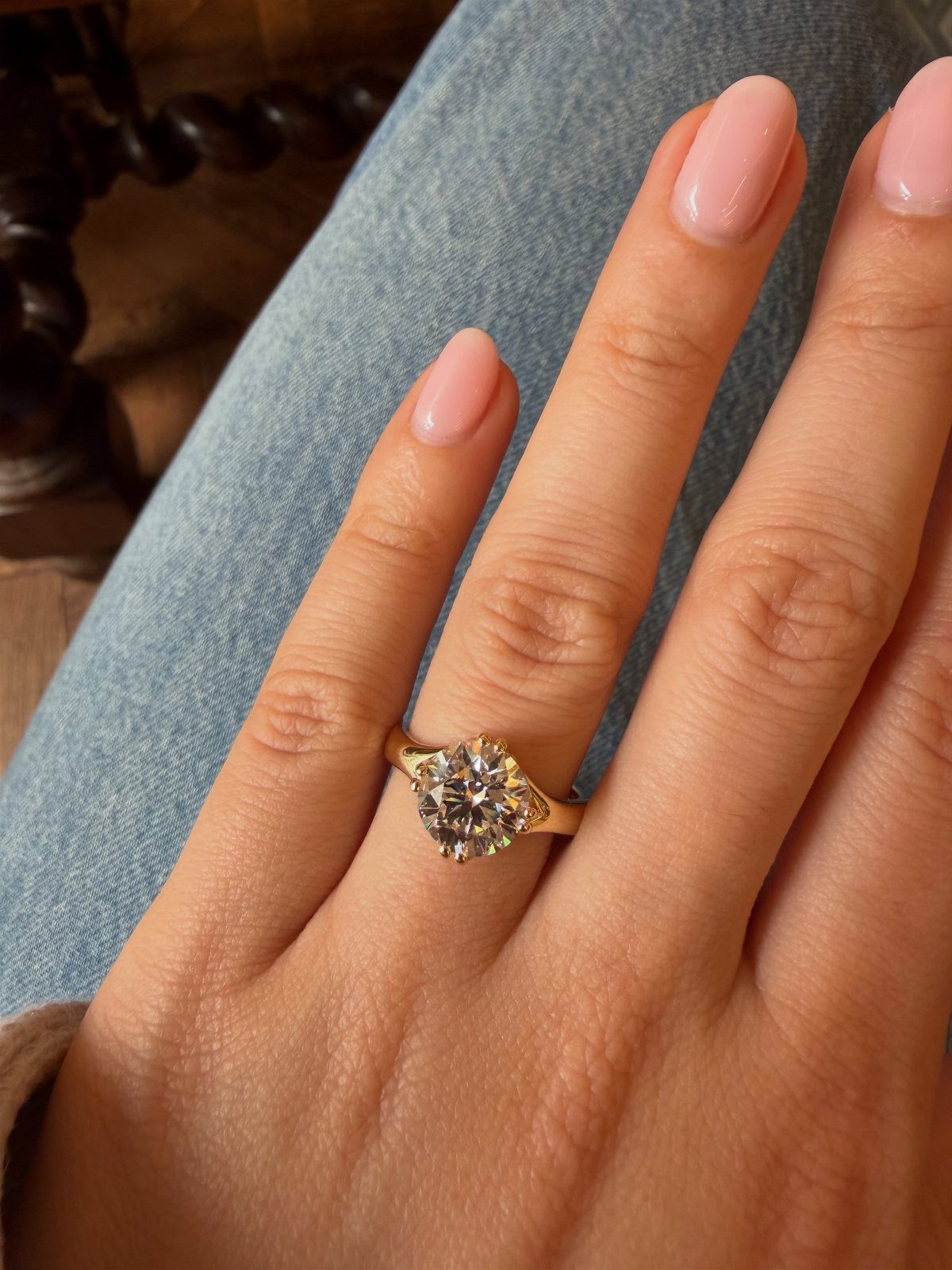 Gold engagement ring with a round diamond on a finger with pink nails