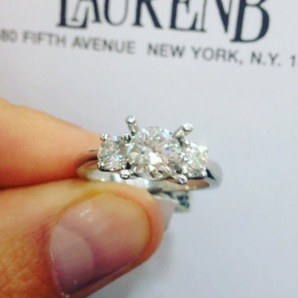 Three-stone diamond engagement ring held by a person against a white background.