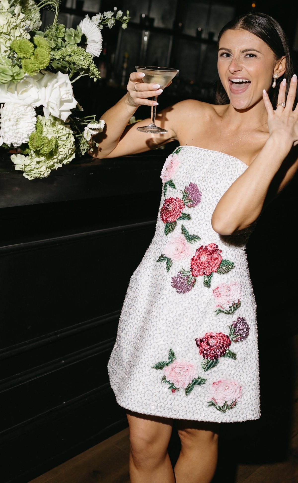 Woman in a short white floral embroidered wedding dress holding a cocktail glass.
