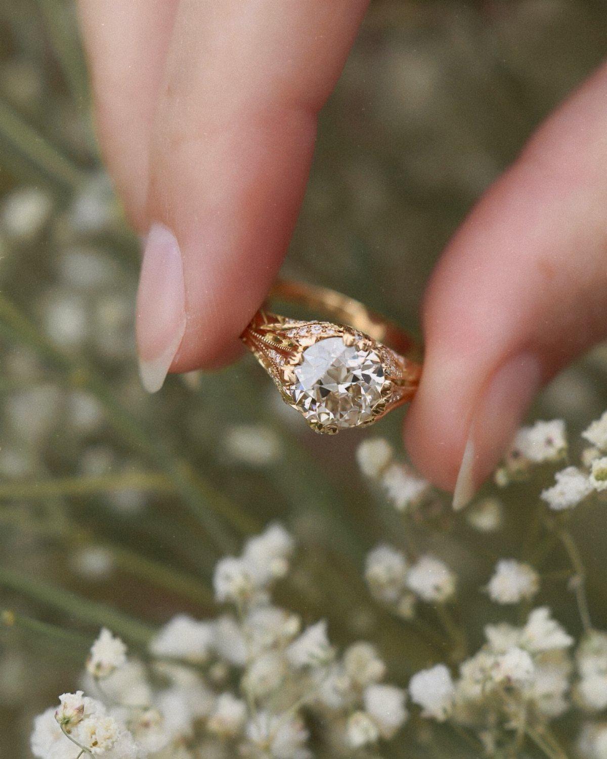 Gold engagement ring with intricate hand-carved settings held against white baby's breath flowers.