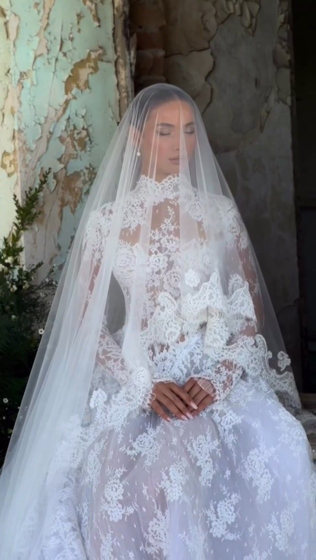 Woman in a voluminous white lace wedding dress and sheer veil against weathered wall