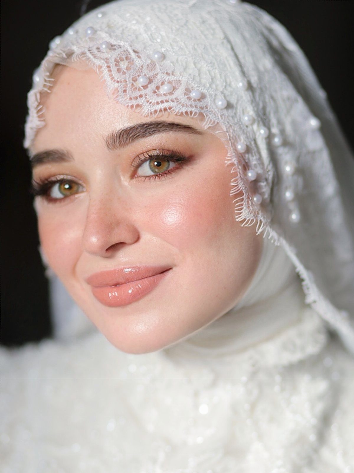 Woman with green eyes wearing bridal makeup and a white pearl-embellished lace hijab.