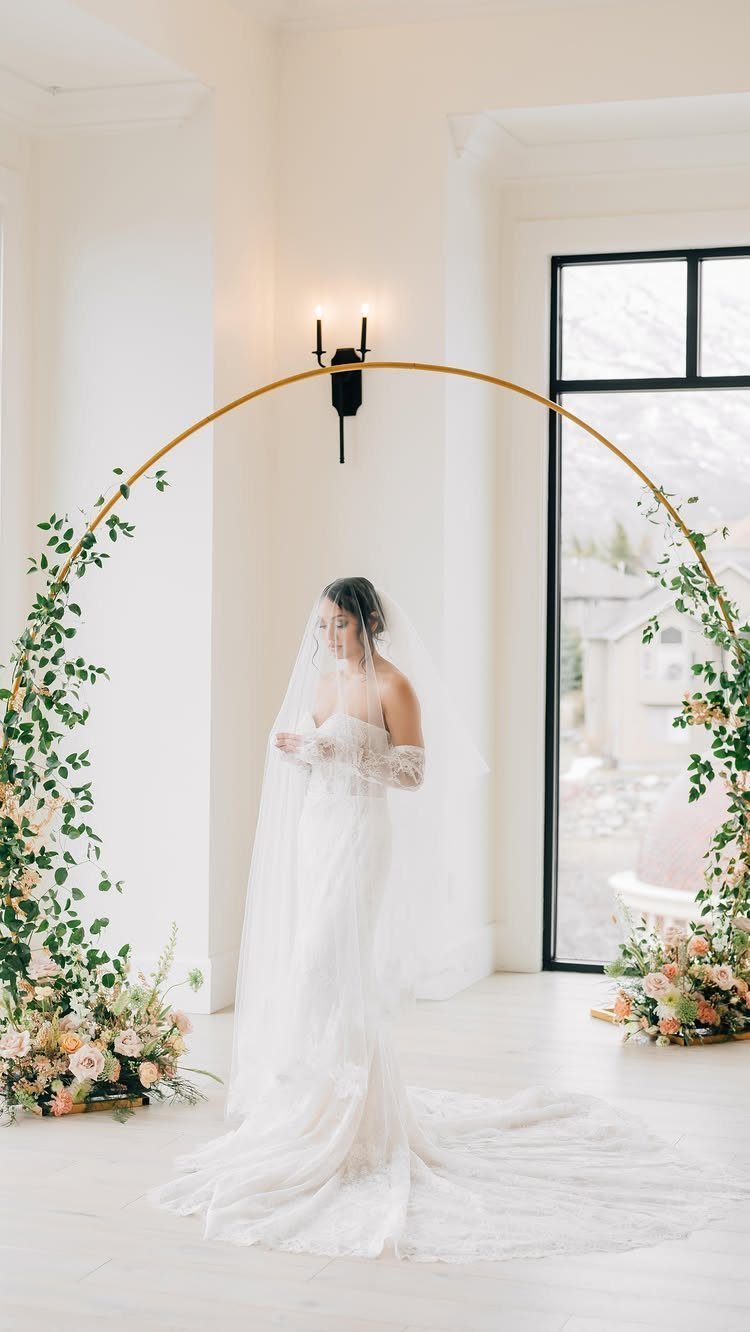 Bride in white gown and long veil standing under a floral gold arch.