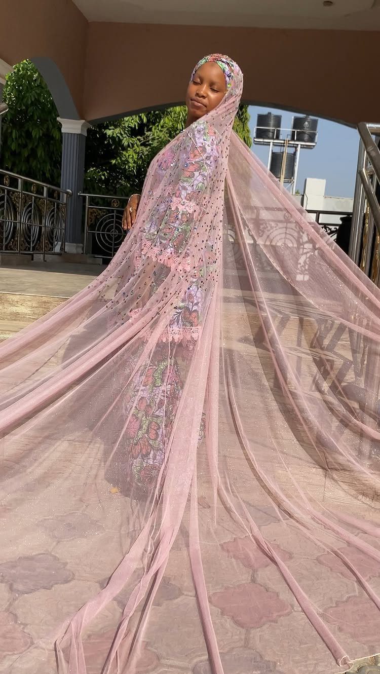 Woman wearing a long, sheer pink embroidered bridal veil and matching headpiece outdoors.