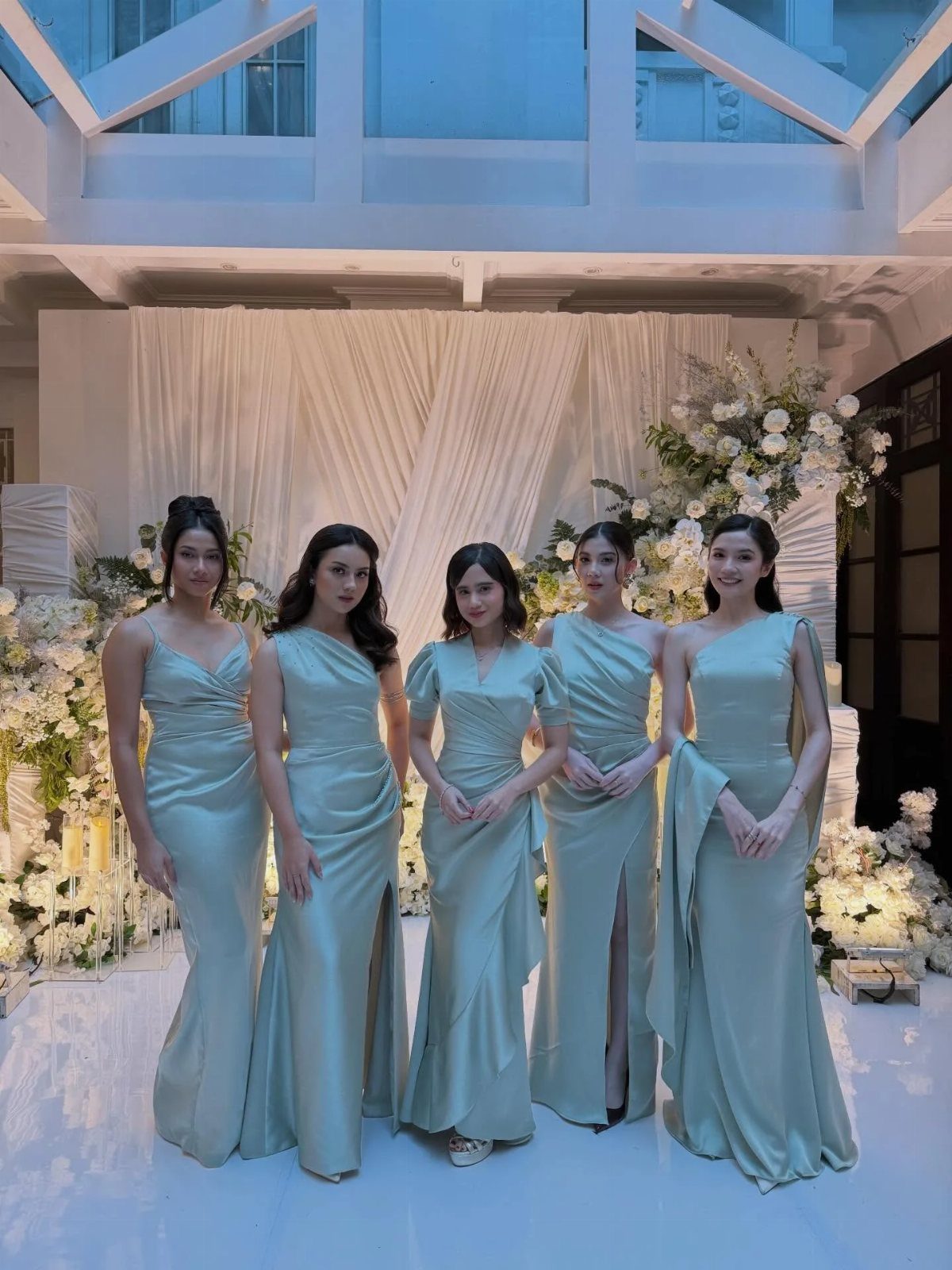 Five women wearing different styles of sage green satin bridesmaid dresses.
