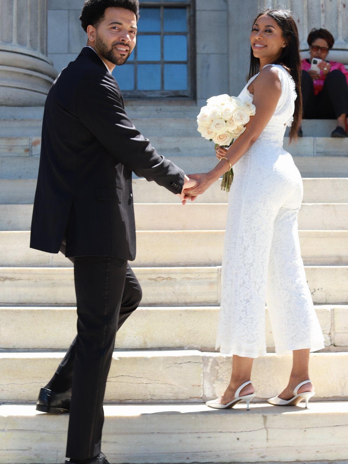 Couple holding hands on stone steps, woman wearing a white lace bridal jumpsuit