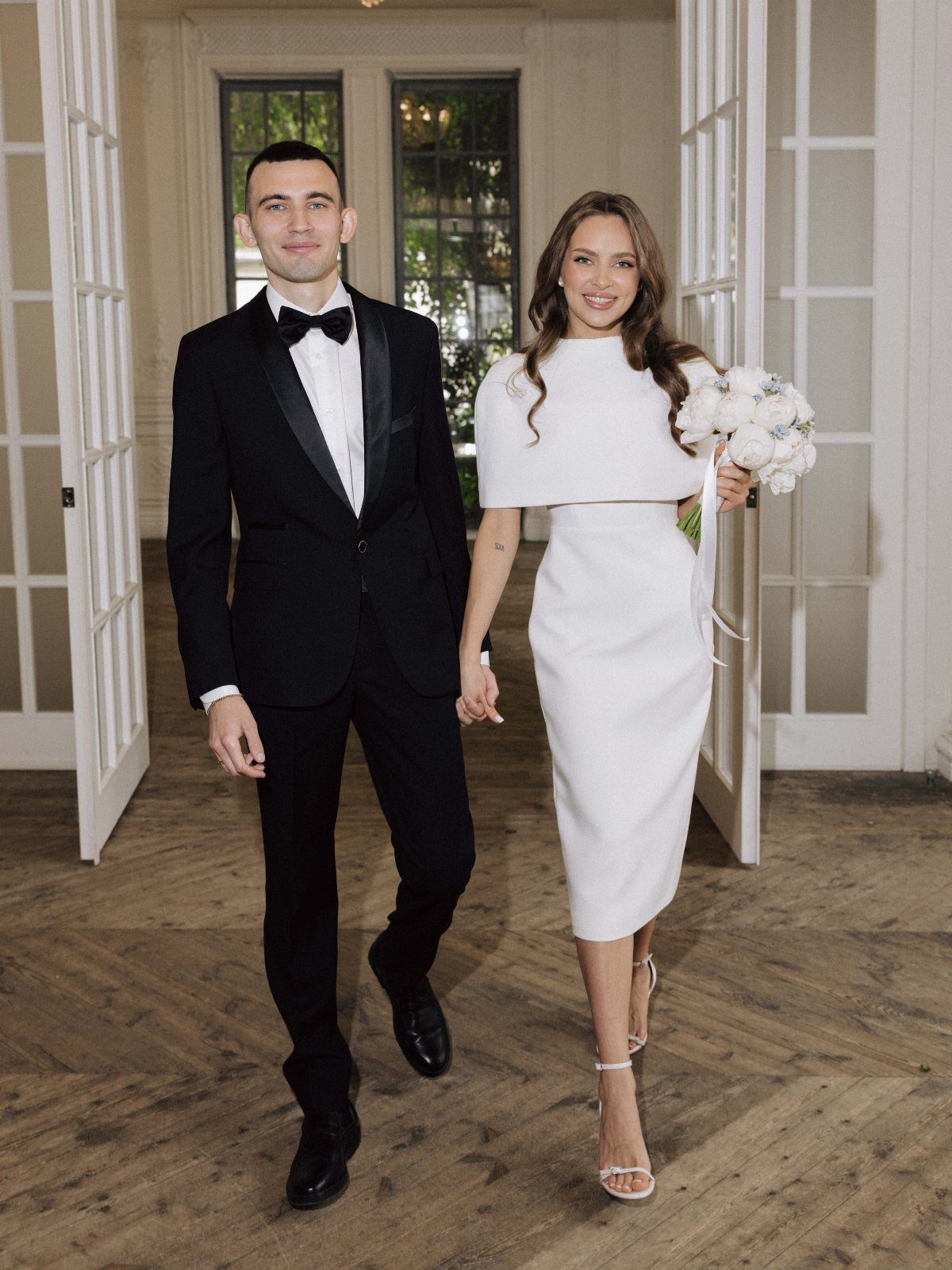 Couple walking hand-in-hand, woman wearing a chic white midi wedding dress with capelet.