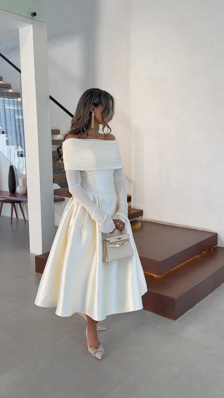 Woman in a white off-the-shoulder midi wedding dress holding a small white handbag.