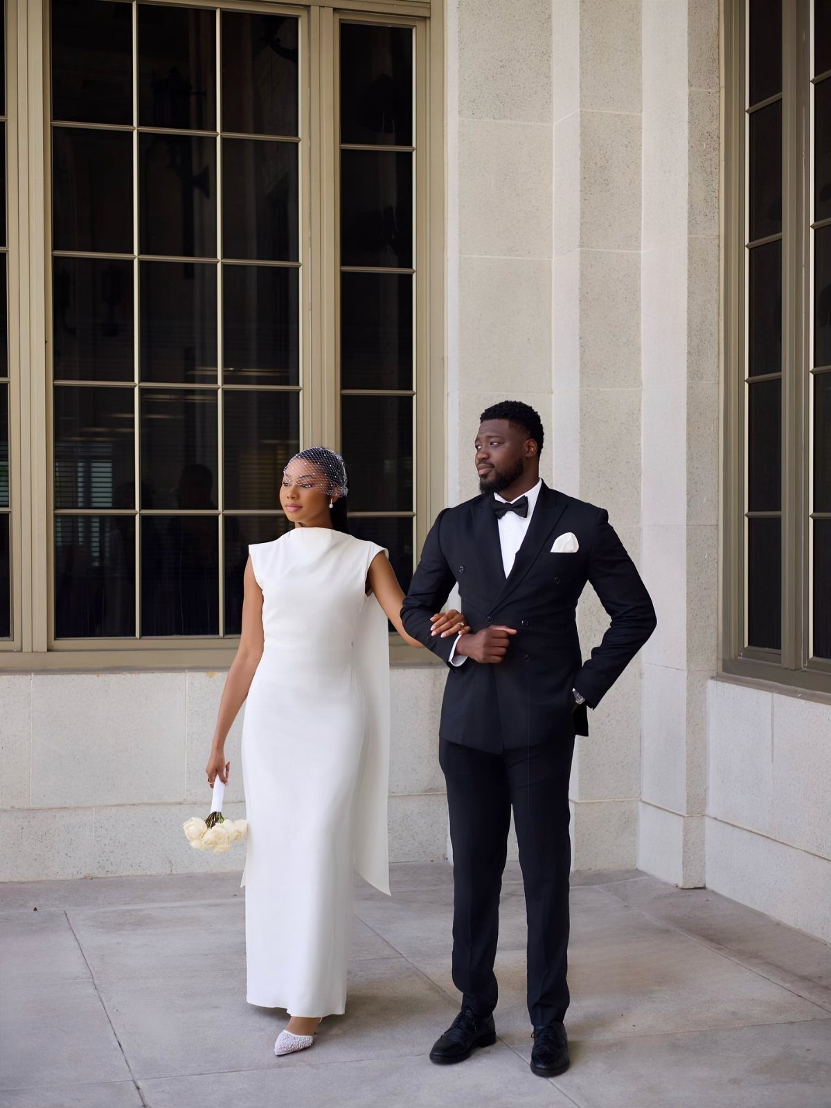 Couple posing outdoors, woman in a sleek white midi dress and man in tuxedo.