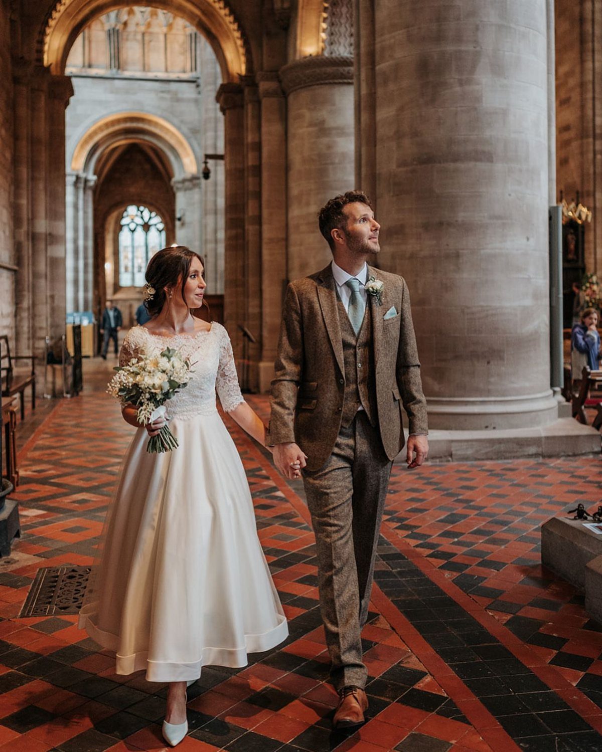 Bride in a white tea-length wedding dress and groom in tweed suit walking through cathedral