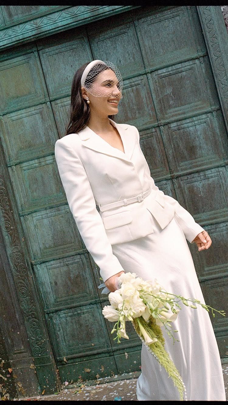 Woman in a white bridal suit and birdcage veil holding white flowers.