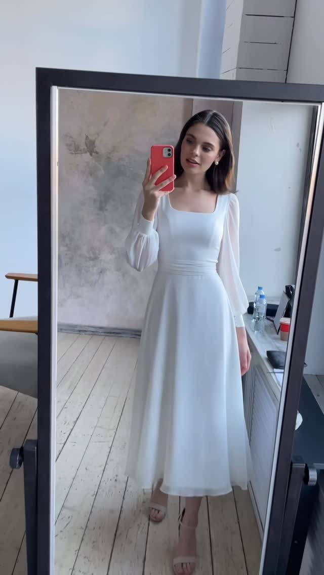 Woman in a white tea-length wedding dress taking a mirror selfie in a room.