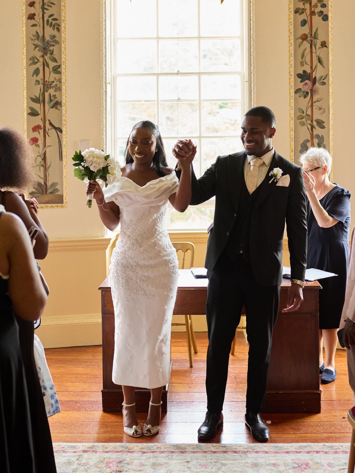 Smiling couple holding hands, woman in off-the-shoulder white midi dress with bouquet.