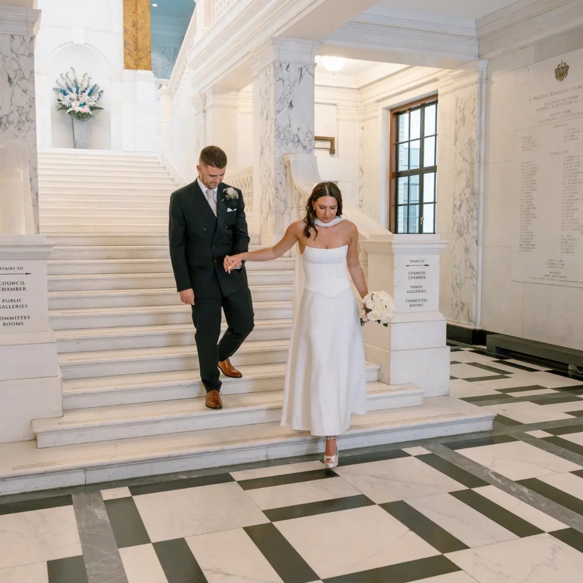 Bride in white A-line dress and groom in suit walking down marble stairs