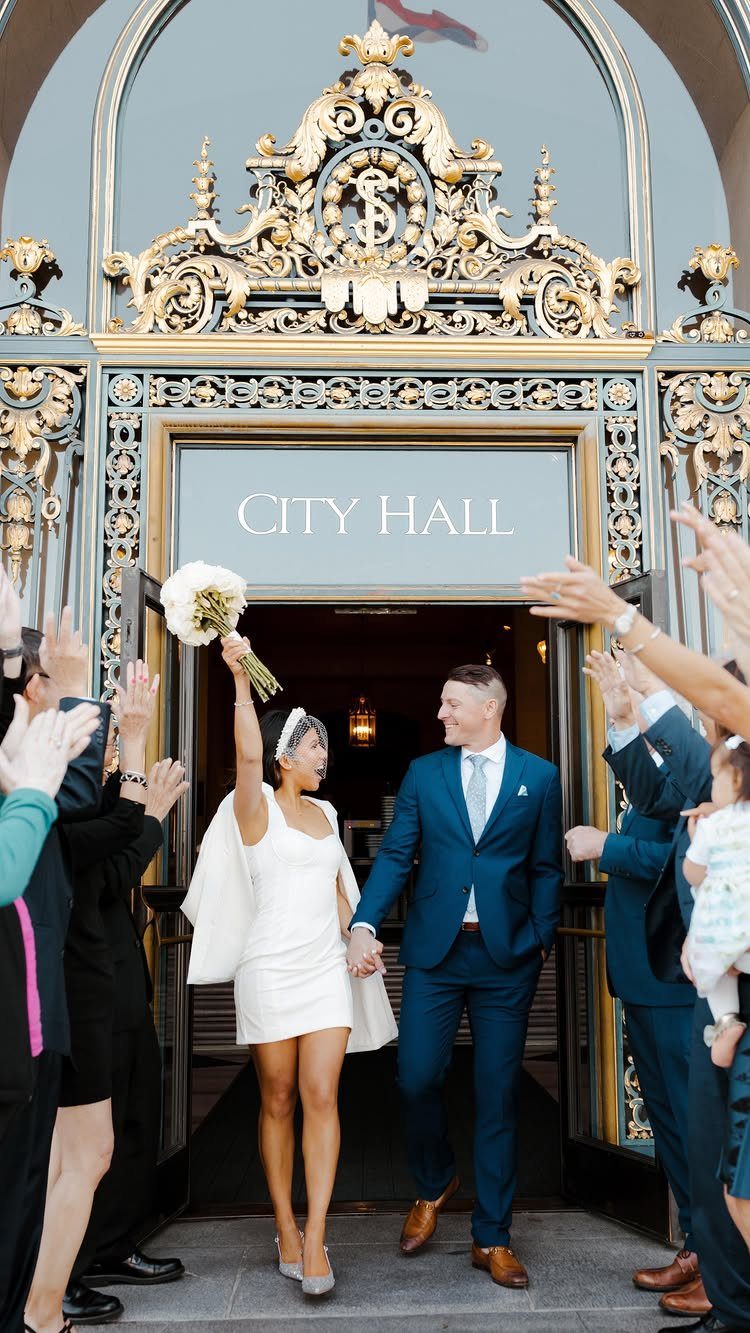 Bride in a short white wedding dress and groom in blue suit exiting City Hall.