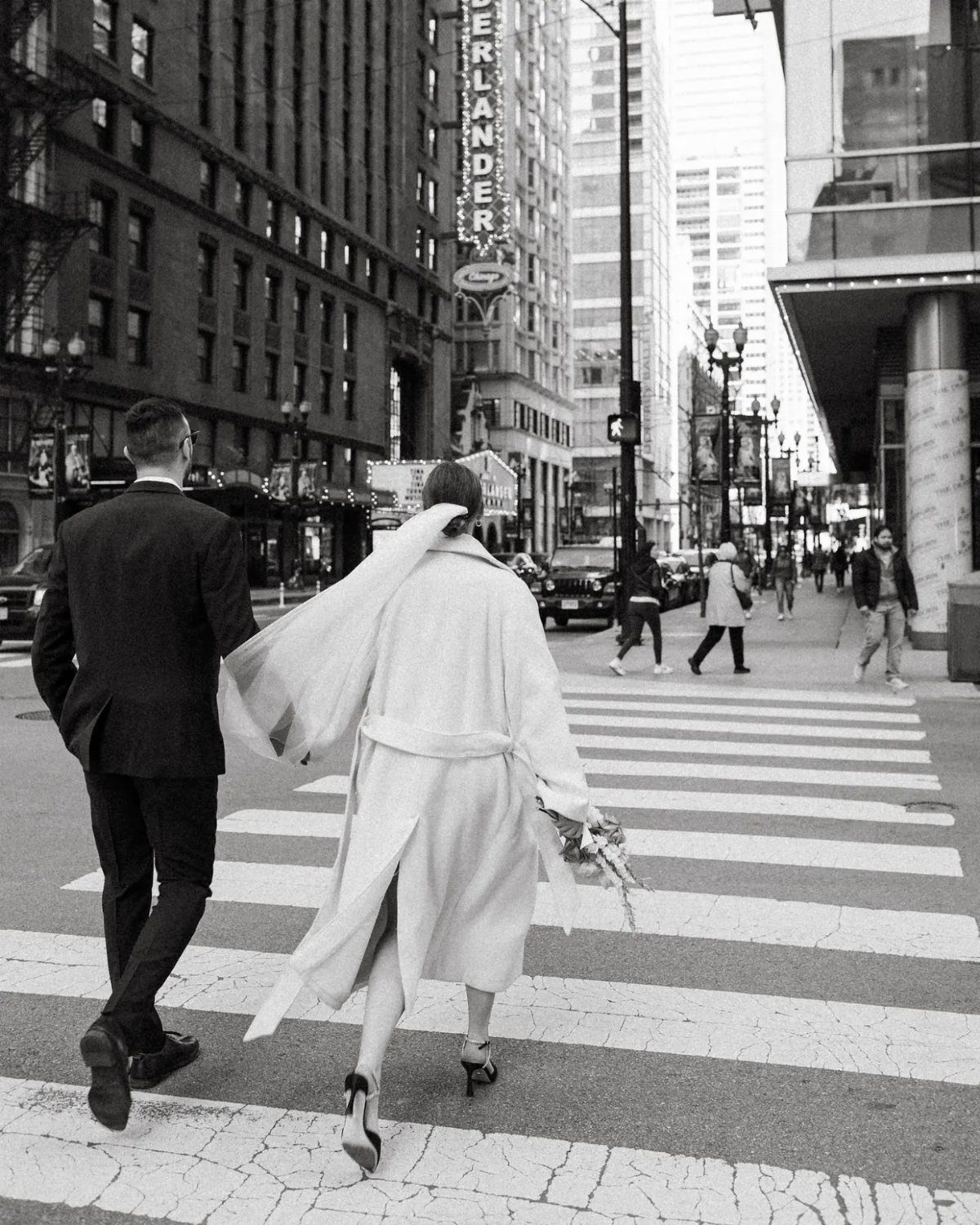 Couple walking across city street, woman wearing a long light-colored bridal coat.