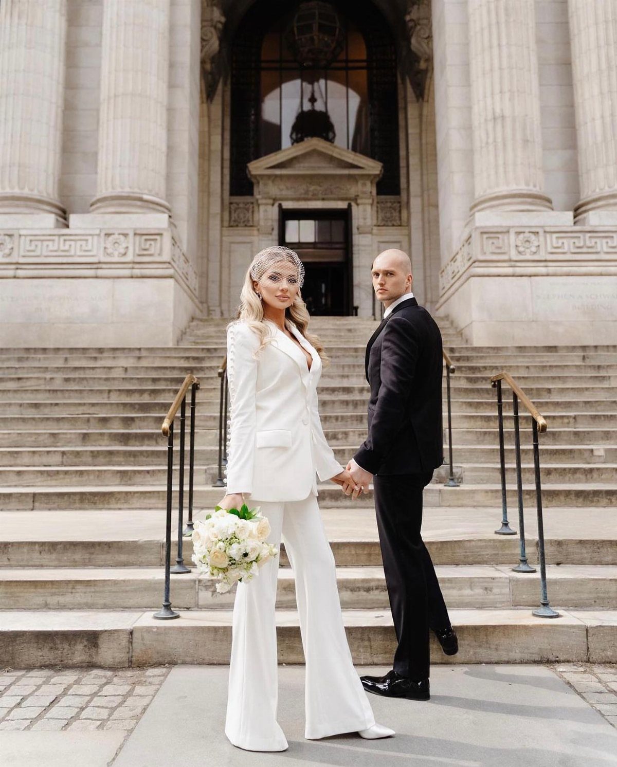 Woman in white wide-leg pant suit and blazer holding bouquet with groom.