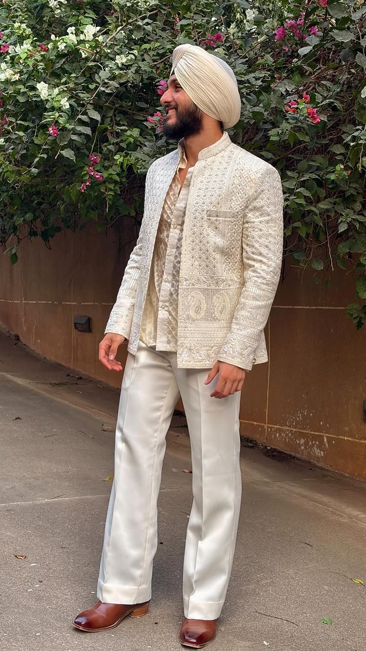 Man in ivory embroidered sherwani and white turban posing outdoors by green bushes.