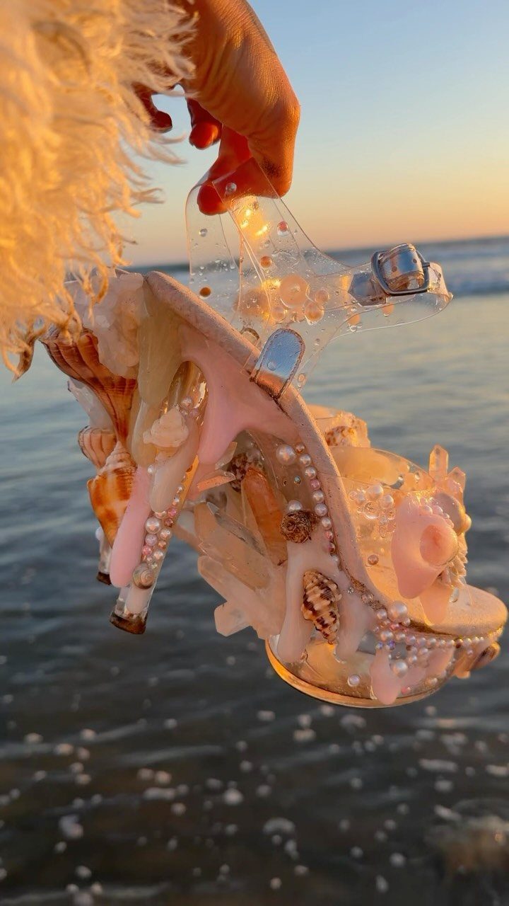 High-heeled sandal decorated with seashells and pearls held against a beach sunset.