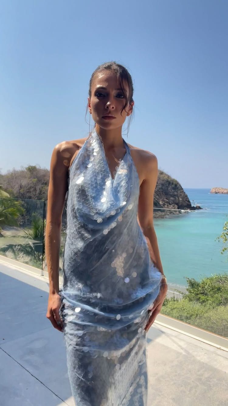Woman in a shimmering blue sequined halter dress overlooking a tropical beach coastline.