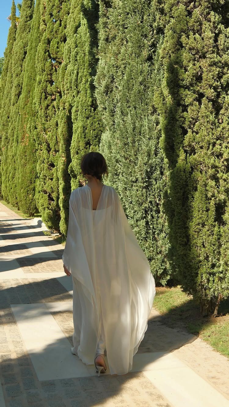 Woman in a white flowy wedding dress walking past tall green cypress trees.