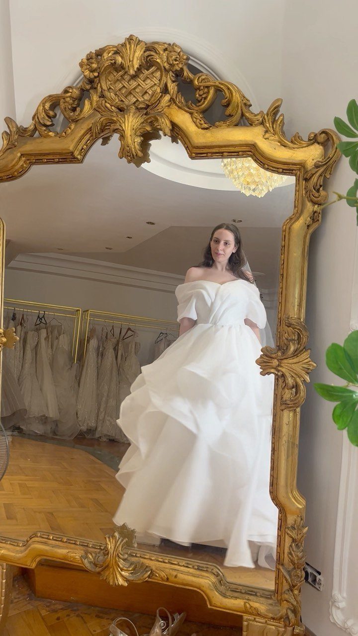 Woman in a voluminous white off-the-shoulder wedding dress reflected in an ornate gold mirror.