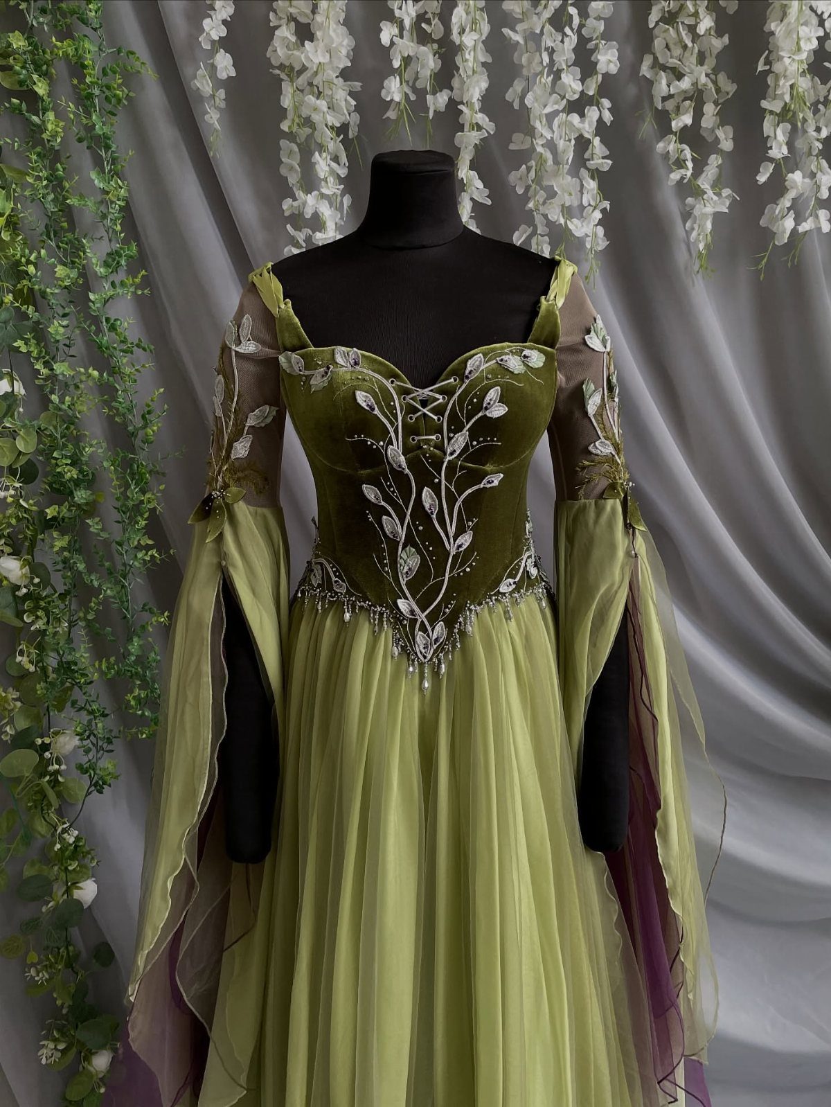 Green gown with silver leaf embroidery on a mannequin against floral backdrop