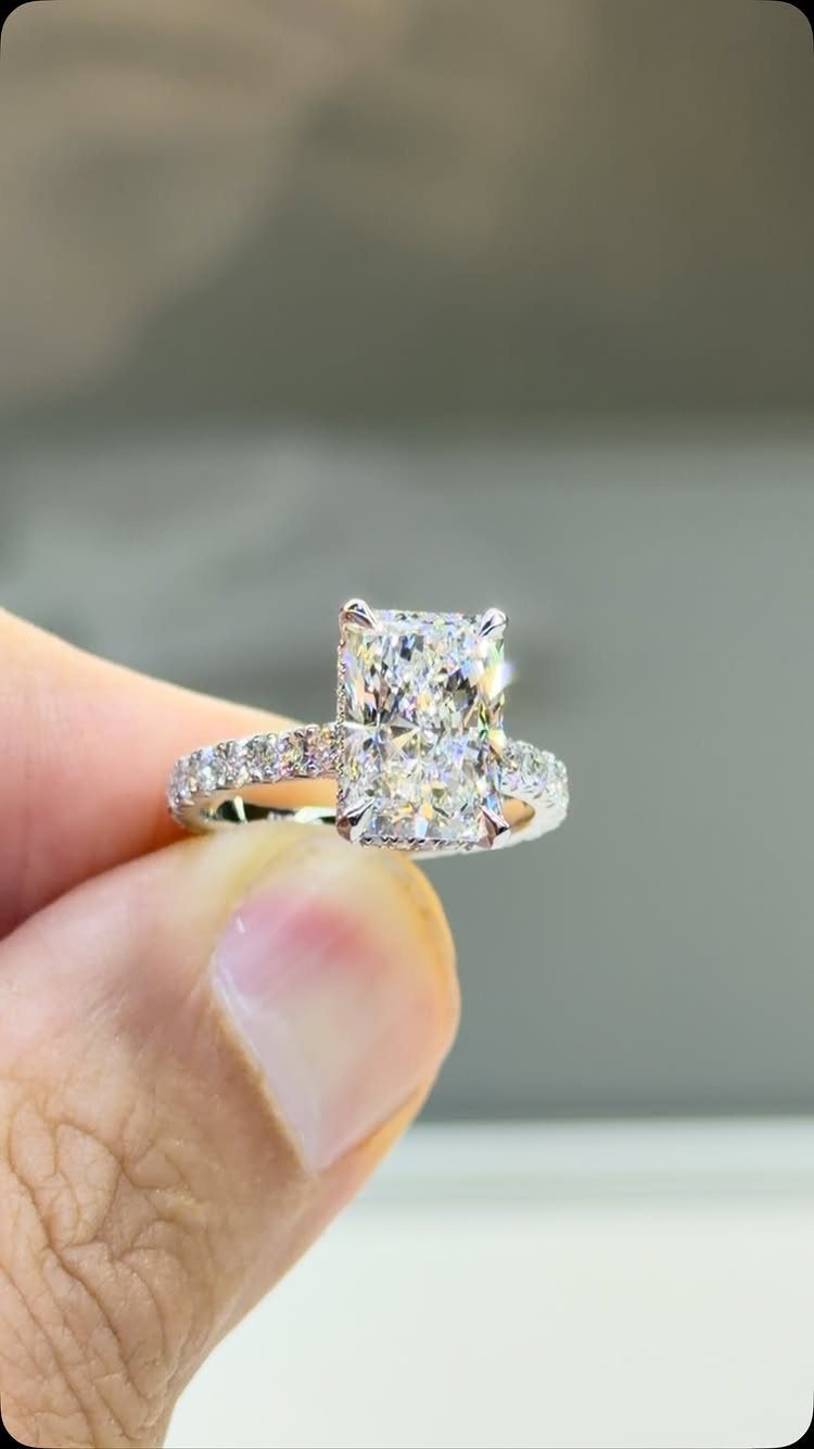 Radiant-cut diamond engagement ring with a paved band held by a finger.