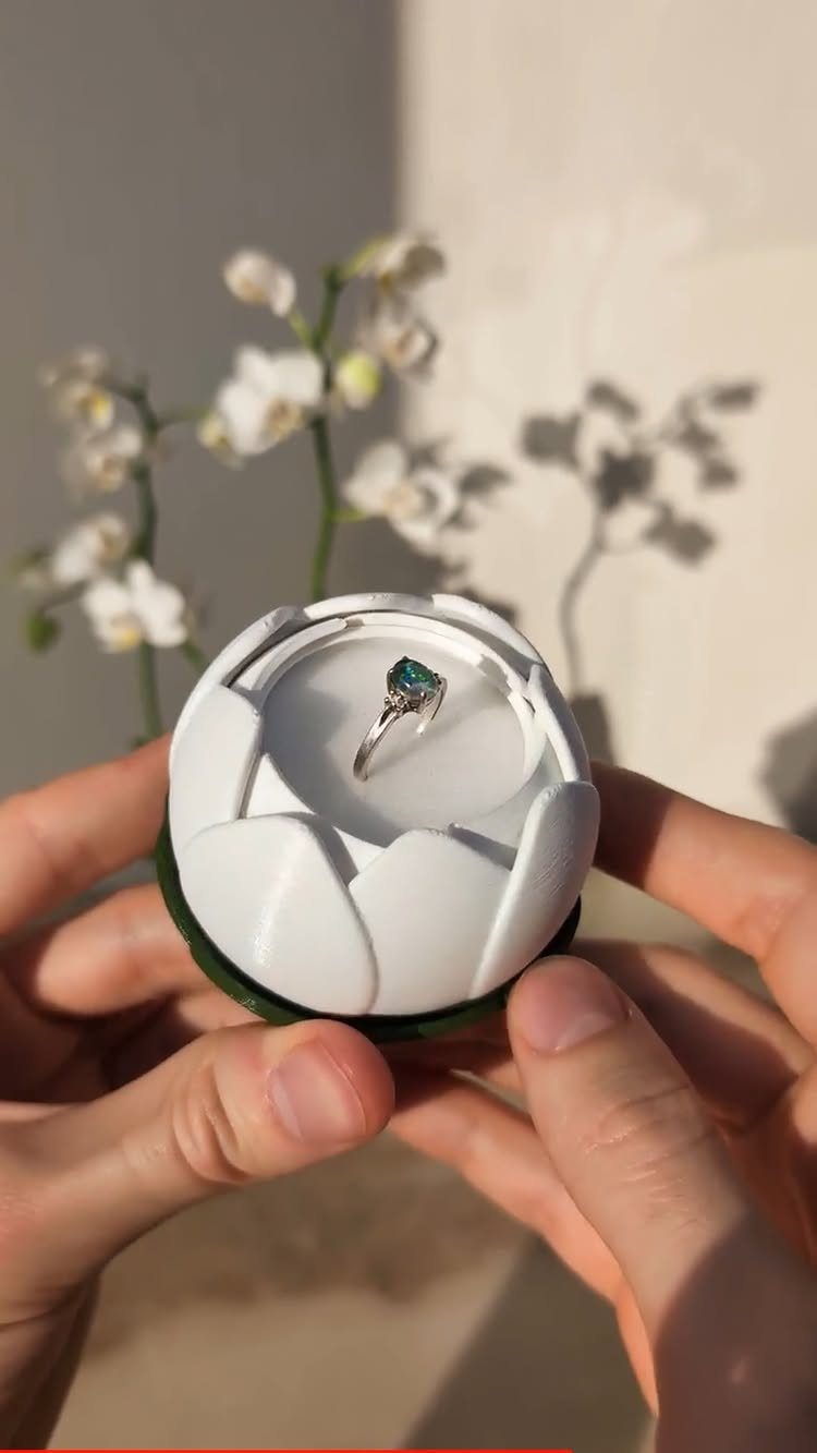 White lotus-shaped 3D printed ring box holding a silver ring with blue gemstone