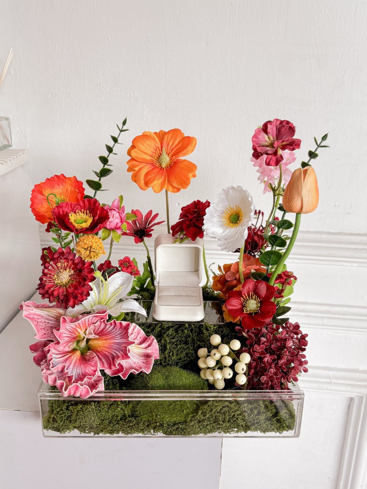 Clear acrylic box with colorful flowers and moss surrounding a white ring box.