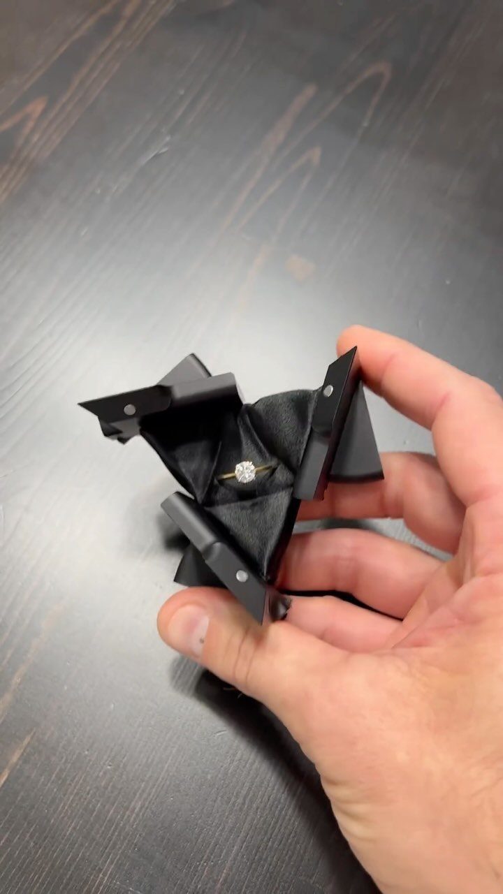 Black geometric origami ring box held in a hand revealing a diamond ring