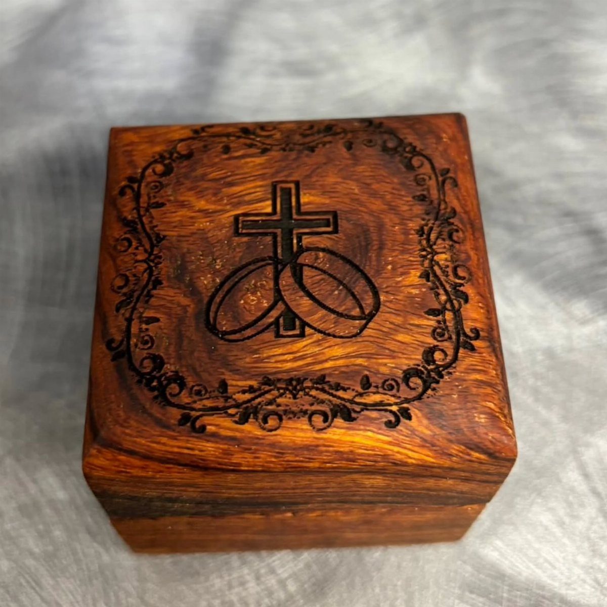 Wooden ring box with engraved cross, interlocking rings, and ornate floral border.