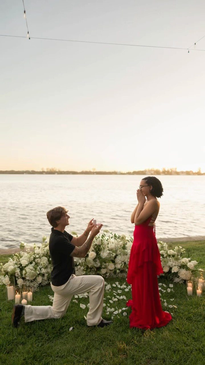 Man proposing to a woman in a red dress by a lake at sunset
