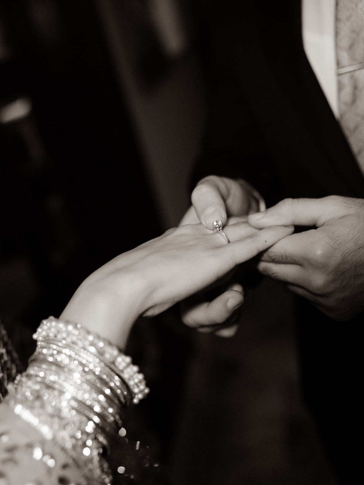 Hands placing a diamond engagement ring on a finger with sparkling bangles.