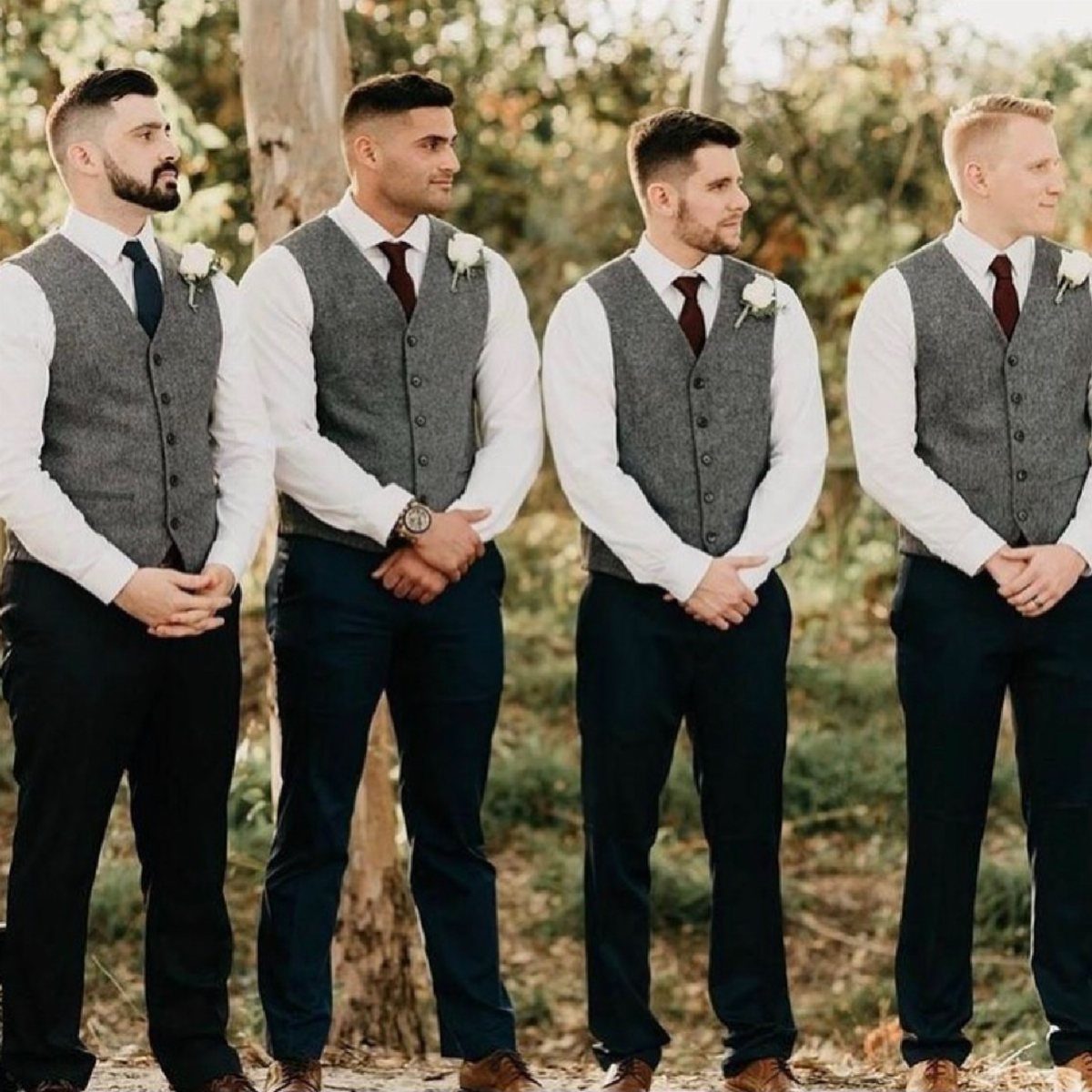 Four groomsmen wearing grey waistcoats, white shirts, and navy trousers in a wooded setting.
