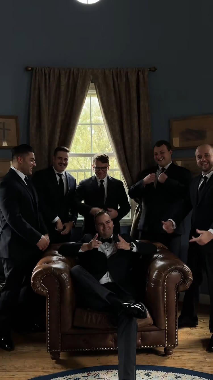 Six men wearing matching black suits and ties posing in a living room.