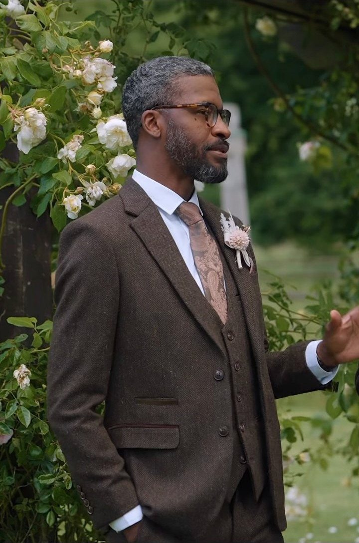 Man in a brown tweed three-piece suit standing beside white roses outdoors.