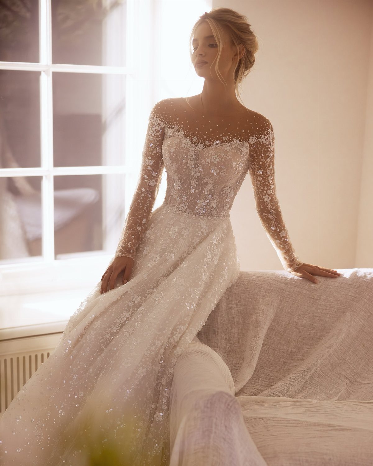 Woman in a shimmering white long sleeve beaded wedding dress posing by a window