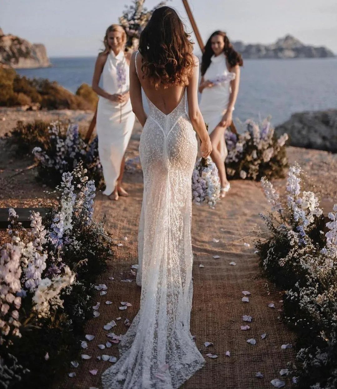 Woman in a white lace sheath wedding dress walking on a floral beach aisle