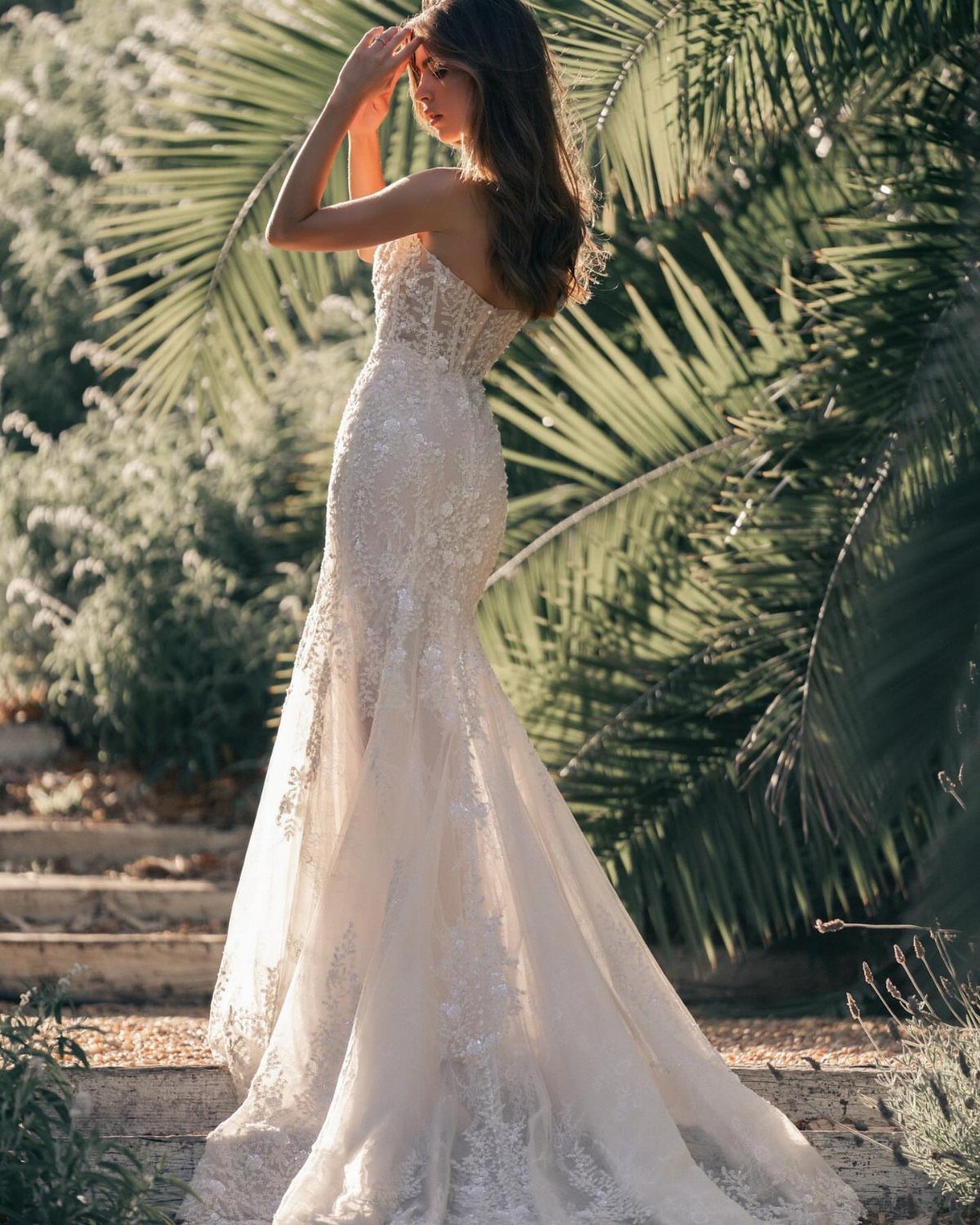 Woman in a strapless white lace wedding dress posing among green palm leaves.