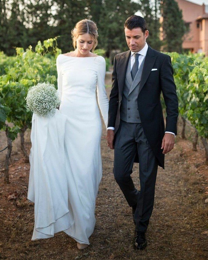 Bride in a minimalist long-sleeved white gown walking with groom in a vineyard