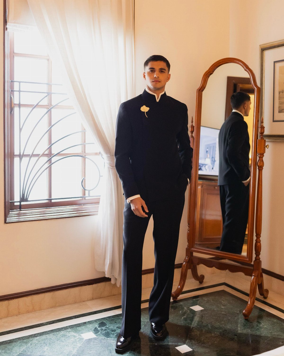 Man wearing a black bandhgala suit and dress shoes standing by a mirror.