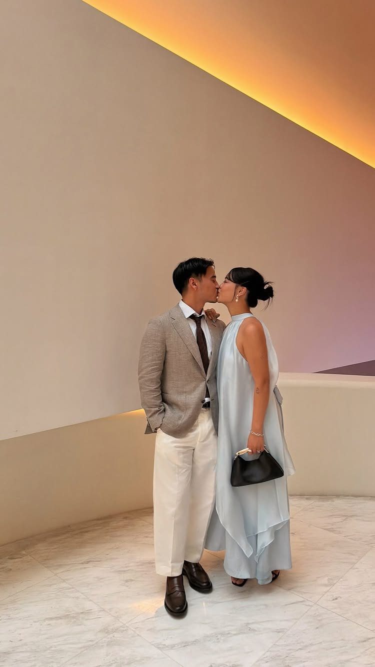 Man in grey blazer and white trousers kissing woman in light blue dress.