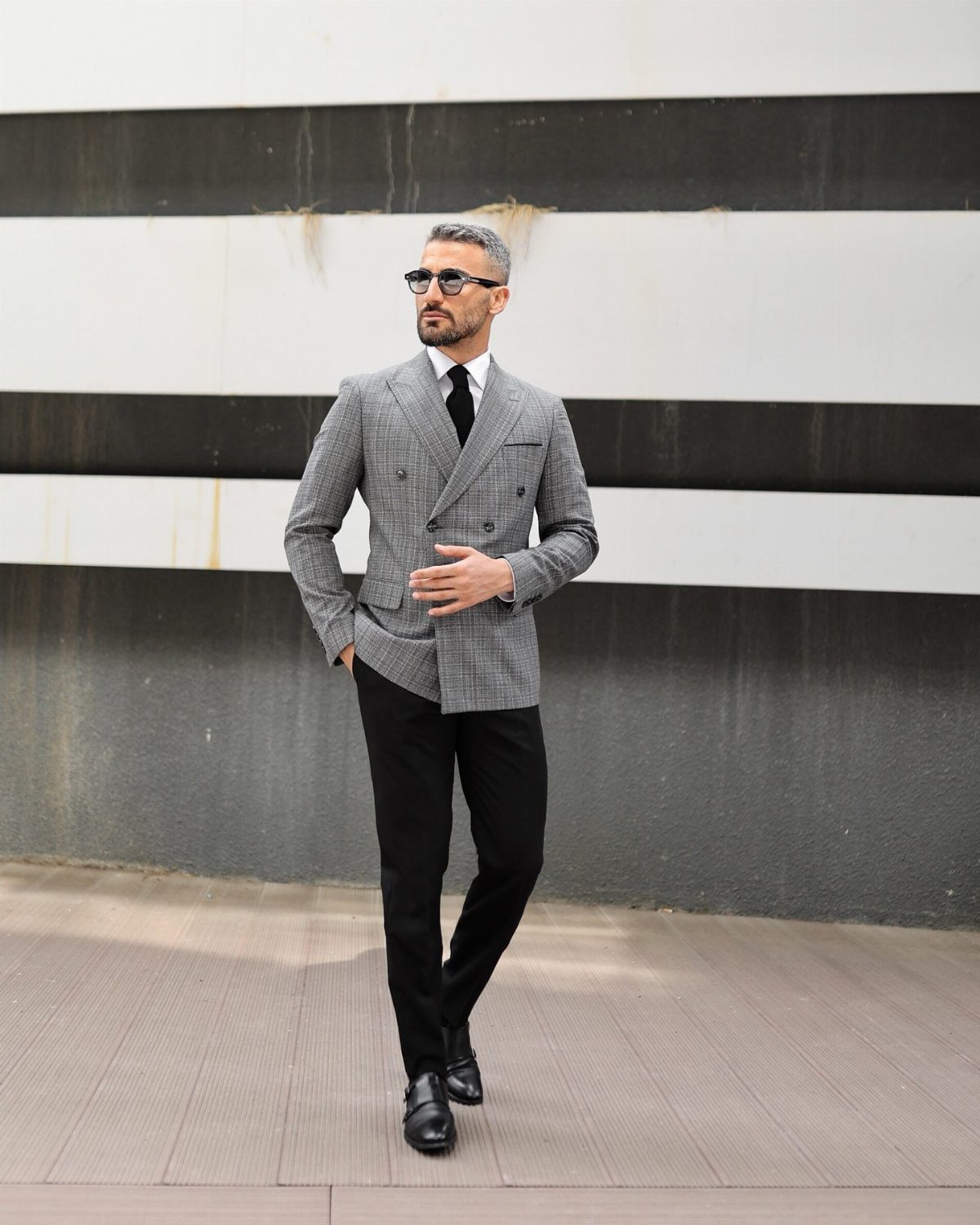 Man in grey plaid blazer and black trousers posing against a striped wall