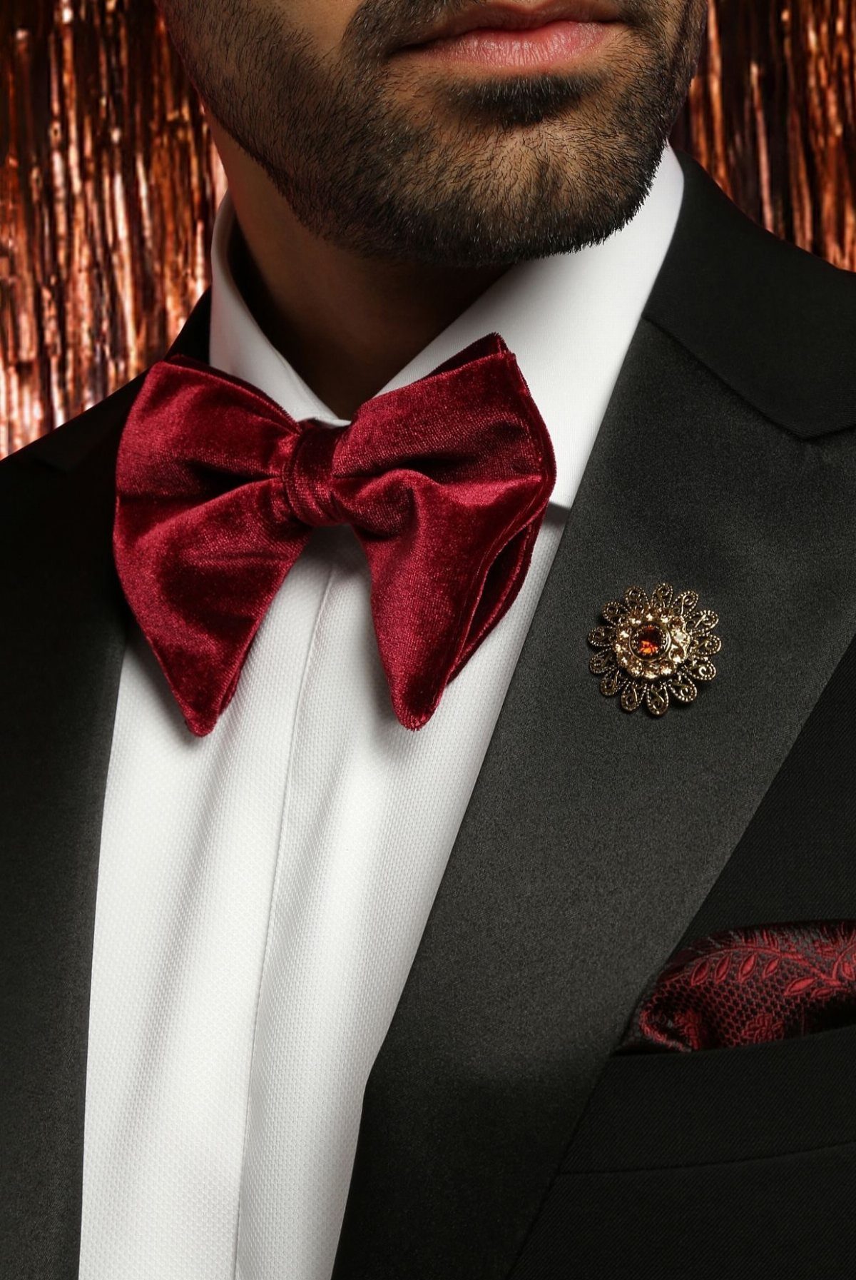 Man wearing a velvet burgundy bow tie with a black tuxedo and gold brooch.
