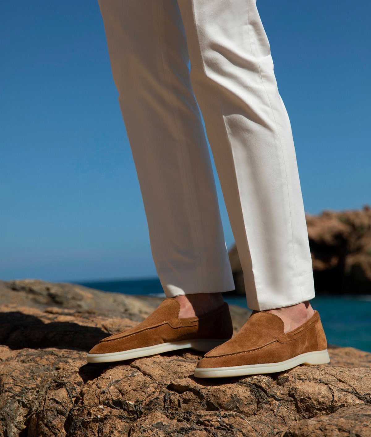 White trousers and brown suede loafers on a rocky coast by the ocean.