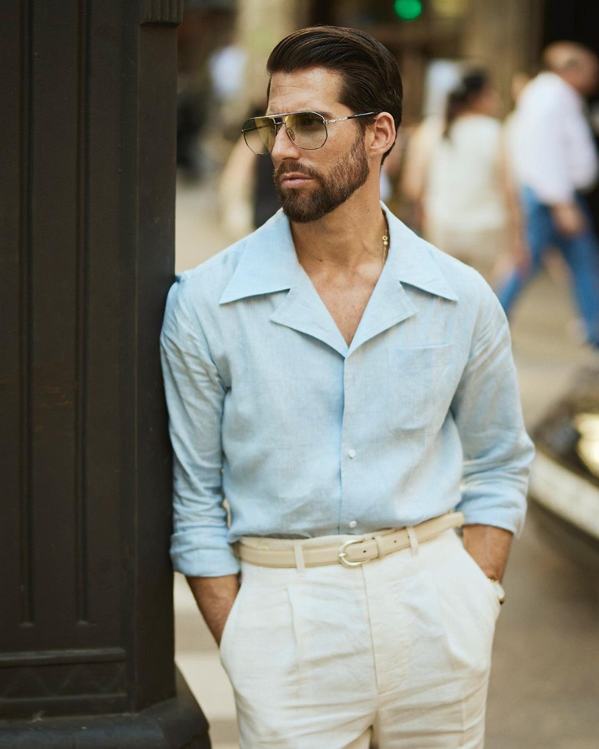 Man wearing a light blue shirt and white trousers leaning against a pillar.