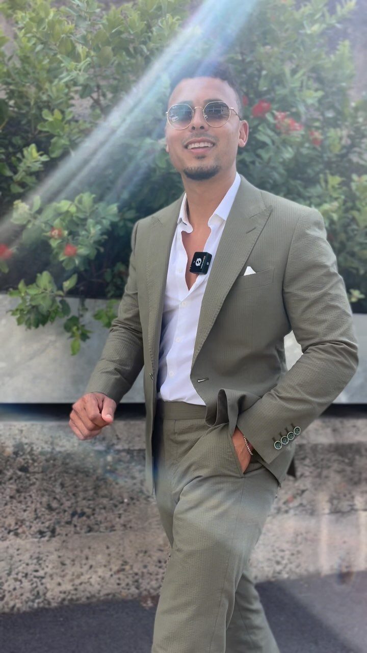 Man in olive green suit and white shirt posing outdoors by green bushes.