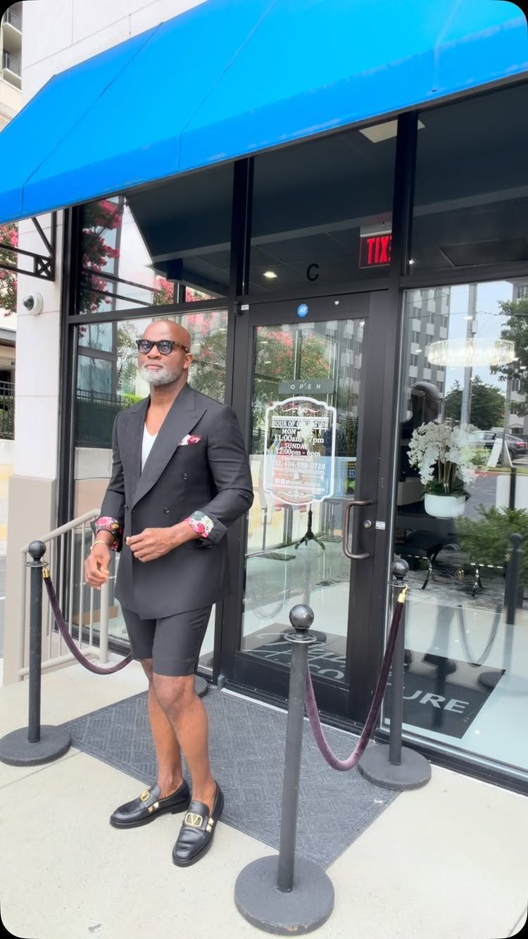Man wearing a black short suit with loafers standing before a blue awning.