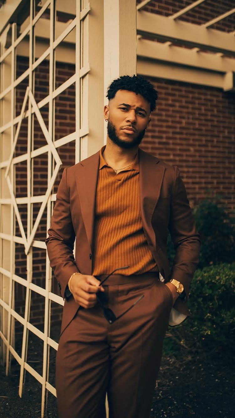Man in a brown suit and ribbed polo shirt posing by a trellis.